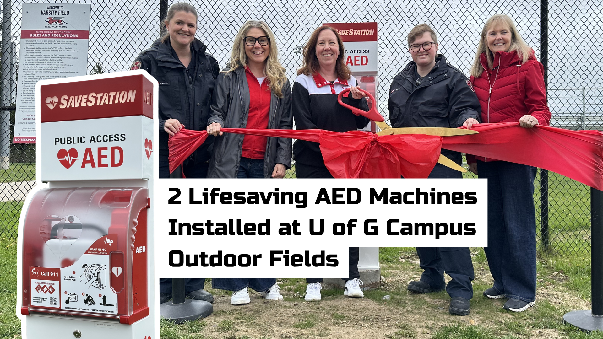People standing in front of a chain-link fence with a public access AED machine. They are holding a large red ribbon tied in a bow. A sign reads, "2 Lifesaving AED Machines Installed at U of G Campus Outdoor Fields.